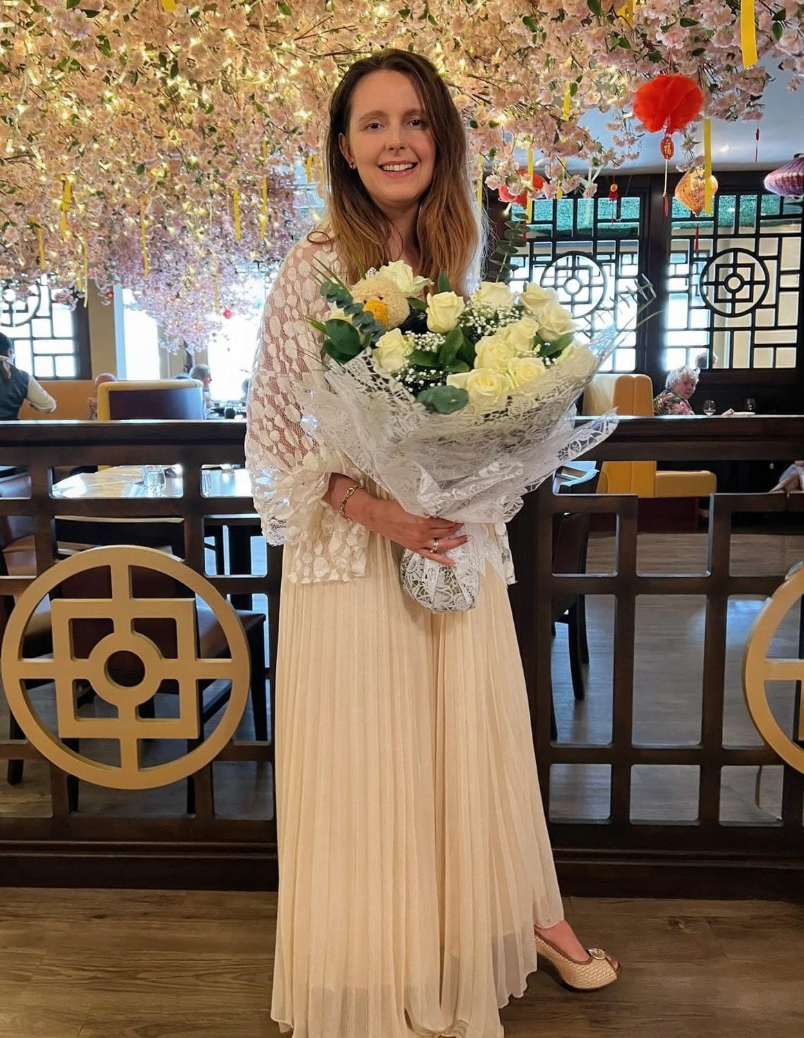 Ewelina (55), liebevolle Polin, hält einen großen Strauß weißer Rosen in einem festlichen, asiatisch inspirierten Restaurant. Diese charmante Damen aus Osteuropa trägt ein elegantes beiges Plisseekleid und einen Spitzenponcho. Möchten Sie Frauen aus Polen kennenlernen? Ewelina ist kreativ und häuslich, liebt romantische Momente und Geselligkeit.